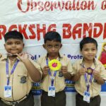 Three schoolboys in beige uniforms pose with handmade badges in front of a raksha bandhan banner each wearing a blue lanyard with an id badge